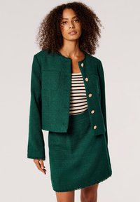Grüne Tweedjacke mit einem cropped Design und goldenen Knöpfen, kombiniert mit einem passenden Minirock. Darunter ist ein gestreiftes Oberteil sichtbar.
