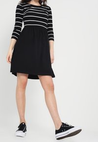 Schwarzes Kleid mit weißen horizontalen Streifen am Oberteil, dreiviertellangen Ärmeln und einem ausgestellten Rock. Kombiniert mit schwarzen Sneakers.