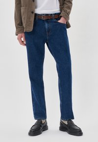 Jeans de mezclilla azul oscuro de corte recto para hombre, con un cinturón de cuero marrón, combinados con mocasines negros y una chaqueta de pana beige.