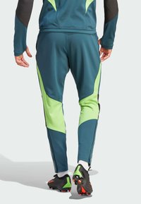 Blauwe sportbroek met groene en zwarte accenten, gemaakt van gladde stof. Heeft taps toelopende benen en ritsen bij de enkels. Draag met zwarte voetbalschoenen.