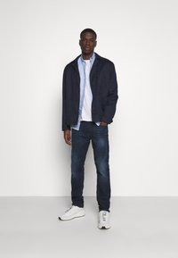 Marinejakke med knapper, lyseblå skjorte under, hvit t-skjorte, mørke jeans og hvite joggesko. Innendørs setting, enkel hvit bakgrunn.