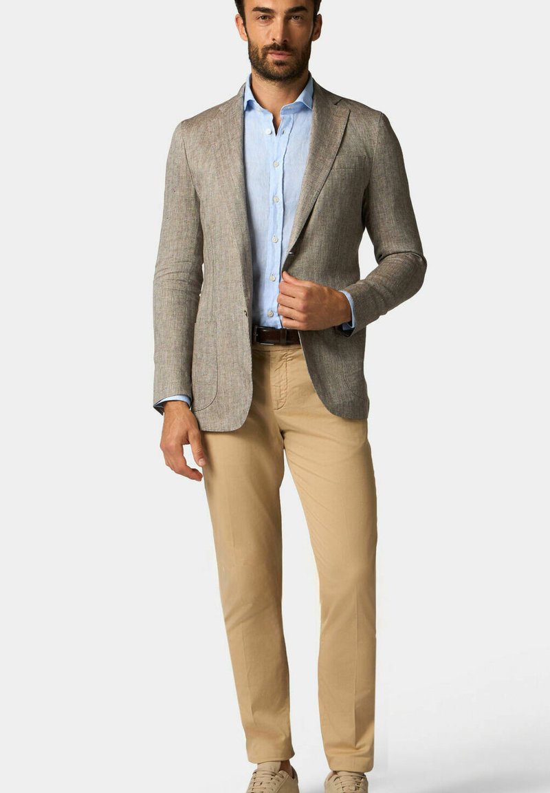 Blazer beige con texture sopra una camicia azzurro chiaro, abbinato a pantaloni khaki chiari e scarpe beige. Vestibilità su misura, due tasche frontali.
