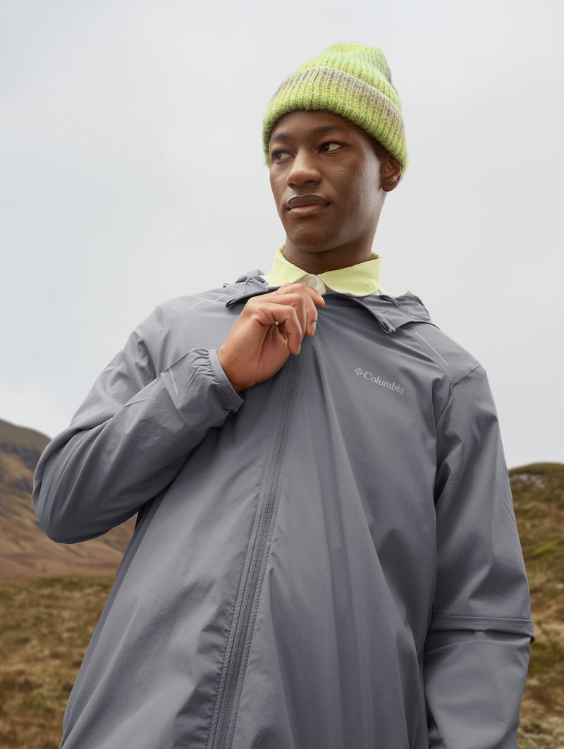 Leichte graue Kapuzenjacke mit Reißverschluss, über einem gelben Hemd getragen. Strickmütze in Grün mit grauen Akzenten. Außeneinstellung.