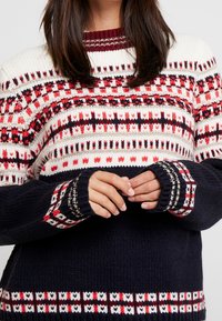 Sweter z dzianiny z kremowym, czerwonym i granatowym wzorem oraz geometrycznymi wzorami. Ściągacze na rękawach i okrągły dekolt dopełniają całość.