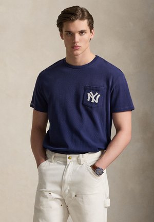 T-shirt bleu marine avec poche avant et logo brodé. Associé à un pantalon beige et une montre. Fond uni mettant en valeur la tenue.