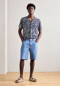 Camisa de manga curta azul-marinho com padrão e design abstrato branco, combinada com calções de ganga azul-claro e sandálias bege. Texturas de tecido suaves.