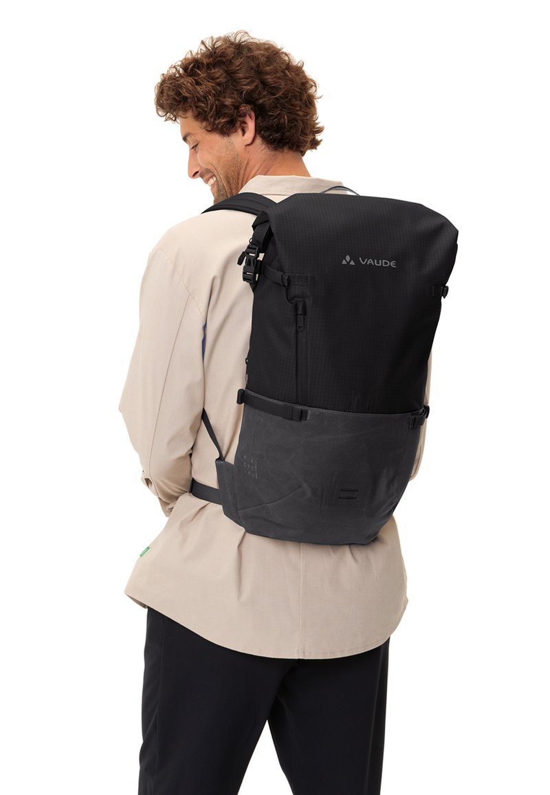 Sac à dos noir avec une surface texturée, des bretelles réglables, une fermeture à rouleau et un branding discret. Panneau arrière dans une teinte plus foncée avec une finition lisse.