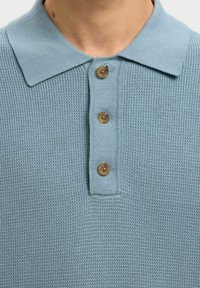 Polo tricoté bleu clair avec trois boutons marron et un col classique porté par une personne.