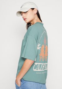 Green cotton t-shirt with oversized fit, featuring a graphic print of two leopards, sun, and palm leaves on the back. Accompanied by a white cap.