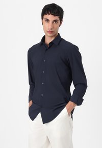 Marineblaue, langärmlige Button-Down-Bluse aus glattem Stoff. Verfügt über einen Standardkragen und ordentlich gefaltete Manschetten, kombiniert mit hellen Hosen.