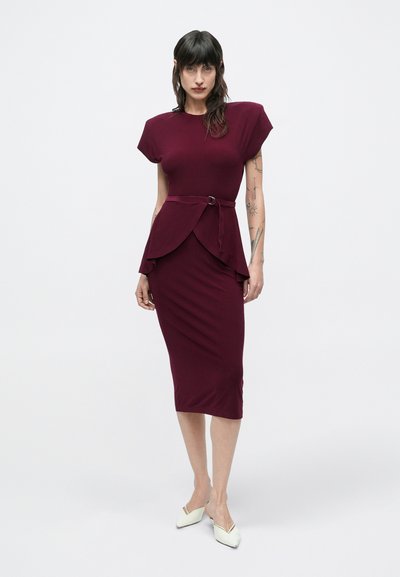 Bordowa dopasowana sukienka z krótkimi rękawami, talią z baskinką i paskiem, zestawiona z białymi butami na cienkim obcasie na jednolitym tle.