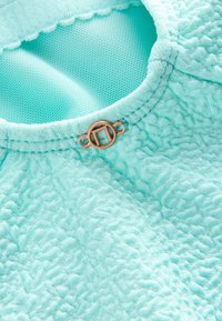 Tissu texturé turquoise clair avec un col arrondi et une petite boucle décorative métallique ronde au col.