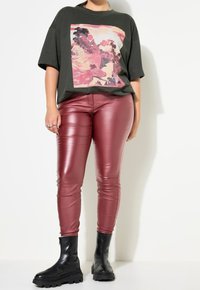 Oversized olijfgroene t-shirt met een roze grafische print, gecombineerd met glanzende bordeauxrode strakke broek en zwarte enkelboots met dikke zolen.