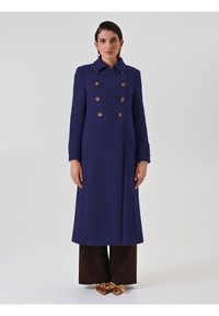 Cappotto doppiopetto blu navy con bottoni dorati, caratterizzato da un ampio colletto e maniche lunghe, abbinato a pantaloni larghi marroni e scarpe metalliche.