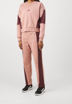 Osoba ubrana w dwukolorowy dres w odcieniach różu i bordo, z pasującą bluzą i spodniami, do tego czarno-białe buty sportowe.