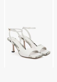 Witte hoge sandalen hebben een gladleren bovenwerk, een open teen ontwerp, dunne enkelbandjes en een slangenprint voetbed met een taps toelopende hak.