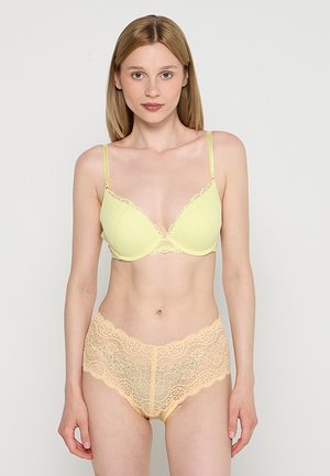 Kobieta o prostych blond włosach, ubrana w jasny żółty biustonosz i beżową koronkową bieliznę, stojąca na jednolitym białym tle.