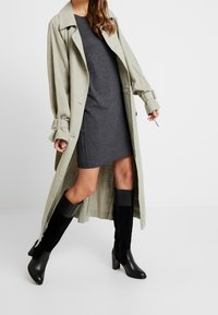Trench-coat long en lin de couleur vert clair, robe mini grise en dessous, et bottes noires montantes en cuir et suede.
