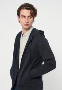 Tmavomodrá mikina na zips s nastaviteľnou kapucňou so šnúrkami, prednými vreckami a zeleným kontrastným prešívaním, nosená cez svetlosivú košeľu.