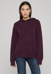 Sweat à capuche couleur prune avec une coupe décontractée, poignets et ourlet côtelés. Tissu texturé à finition douce, associé à un jean bleu.