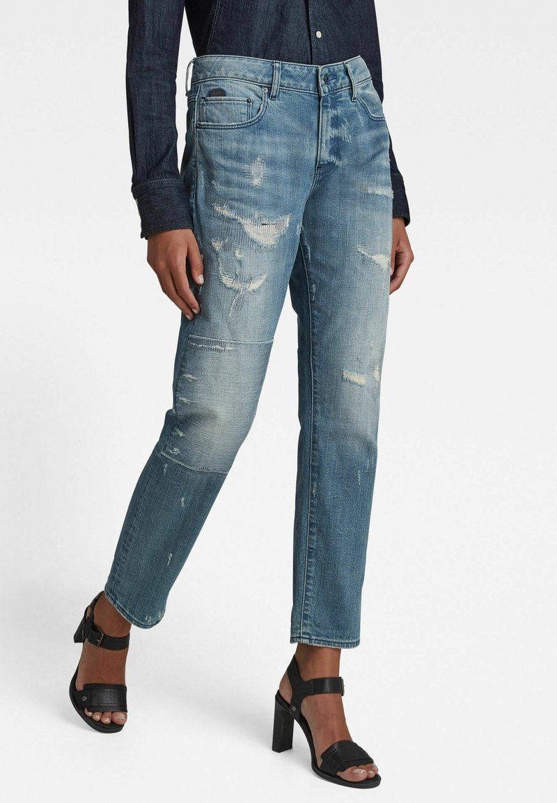 Blue denim jeans with distressed accents, straight-leg cut, and subtle fading. Paired with black heeled sandals, showcasing a casual look.