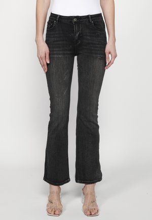 Femme portant un jean noir légèrement évasé avec des détails subtils usés et des sandales transparentes à talons, debout devant un fond uni.