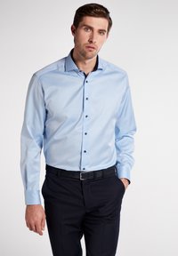 Camicia azzurra chiara a maniche lunghe con colletto alla francese, bottoni neri e texture liscia, indossata con pantaloni scuri e una cintura nera.