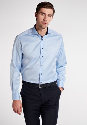 Himmelblaues Hemd mit langen Ärmeln und Button-Down-Kragen, schwarzen Knöpfen und glatter Textur, kombiniert mit dunklen Hosen und einem schwarzen Gürtel.