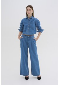 Camisa de mezclilla con botones y un bolsillo en el pecho, combinada con pantalones vaqueros de pierna ancha, ambos en un tono azul claro. Tacones negros de punta.