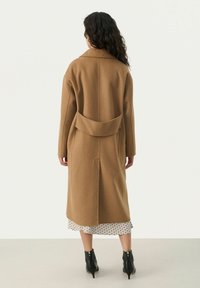 Langer, brauner Wollmantel mit großem Kragen, Rückenschlitz und leicht strukturierten Ärmeln. Trägt über einem weißen gepunkteten Kleid und schwarzen Stiefeln.