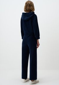 Combinaison en velours bleu marine avec capuche, manches longues et taille élastique. Elle possède des pantalons à jambes larges et des poignets côtelés. Les chaussures sont de couleur claire.