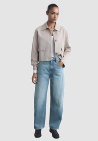 Gekorfde beige jas met een kraag, twee voorkeurszakken en een knoopsluiting, gecombineerd met lichtblauwe wijde jeans en zwarte schoenen.
