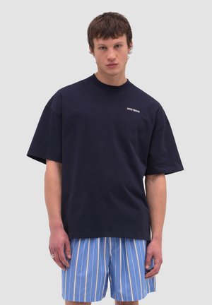 Giovane uomo che indossa una maglietta larga blu scuro con la scritta "WRSTBHVR" e pantaloncini a righe blu, in piedi contro uno sfondo chiaro e semplice.