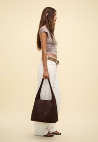 Borsa a spalla in suede marrone con una texture morbida, forma flessibile e una sola maniglia larga, tenuta da una persona che indossa un top aderente e pantaloni bianchi.