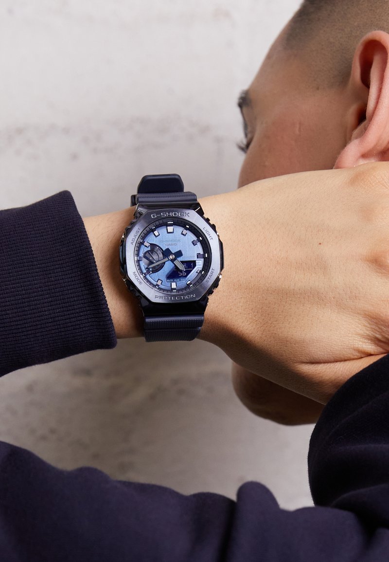 Ceasul de mână G-Shock are un cadran rotund de culoare albastră cu accente argintii, o bratara neagră și mai multe butoane funcționale. Design durabil.