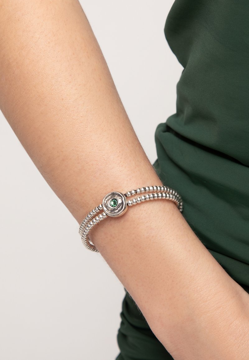 Pulsera de plata con un diseño de cuentas, que presenta una pieza central redonda de plata adornada con una gema verde. Se lleva en la muñeca sobre una prenda verde.