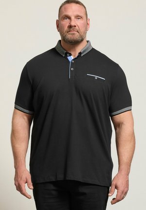 Mężczyzna z brodą ubrany w czarną koszulkę polo z krótkim rękawem, z szarym kołnierzykiem i mankietami, stojący na tle jasnego, jednolitego tła.