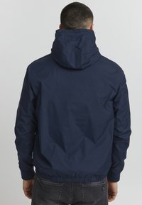 Chaqueta con capucha de color azul marino, fabricada con un material ligero y suave. Presenta puños y cintura elásticos. La vista trasera muestra un diseño simple sin adornos.
