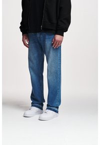 Jeans de mezclilla azul claro, corte relajado, con textura desgastada. Llevados con zapatillas blancas y una sudadera negra con cremallera. Detalles de costura estándar visibles.