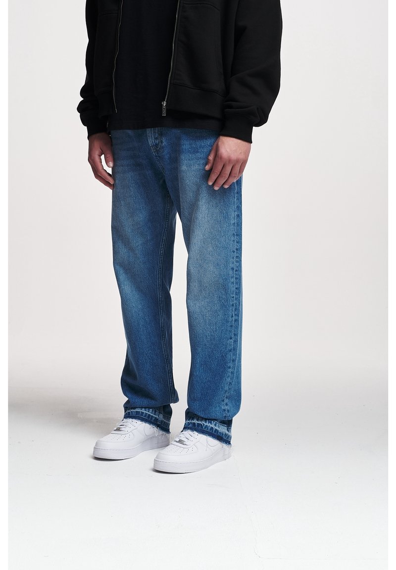 Jeans de mezclilla azul claro, corte relajado, con textura desgastada. Llevados con zapatillas blancas y una sudadera negra con cremallera. Detalles de costura estándar visibles.