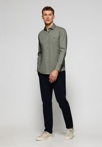 Camicia verde chiaro con colletto a punta, maniche lunghe e un piccolo logo sul petto, abbinata a pantaloni blu navy scuri e scarpe beige.