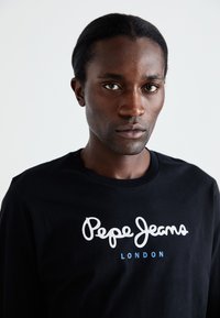 Camisa negra de manga larga con el logo de "Pepe Jeans" en blanco y "LONDON" en azul, hecha de algodón y con un clásico cuello redondo.