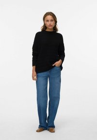 Maglione nero in maglia con texture a coste, collo alto; abbinato a jeans azzurri a gamba larga. Scarpe marroni senza lacci completano il look.