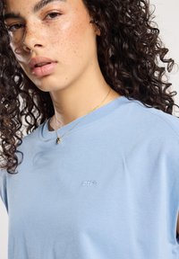 Giovane donna con capelli ricci che indossa una t-shirt azzurra chiara e una piccola collana d'oro con un pendente su uno sfondo neutro.