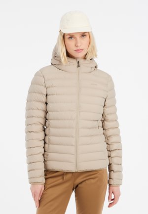 Frau trägt beige wattierte Jacke mit Kapuze, beige Mütze und braune Hose, steht frontal vor einem einfarbigen weißen Hintergrund.
