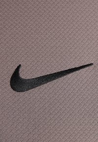 Logo de Nike en negro, bordado en un tejido texturizado de color marrón claro. El diseño presenta una forma de swoosh suave y curvada, enfatizando la identidad de la marca.