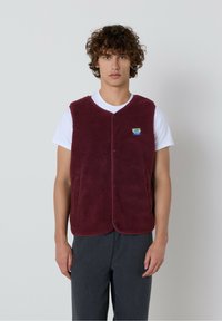 Tumši sarkans polārdzīves vestes izstrādājums ar teksturētu virsmu, noapaļotu apkakli un spiedpogu aizdari, uzvilkts virs baltas T-kreklu. Kreklā ir neliels logotips kreisajā krūtī.