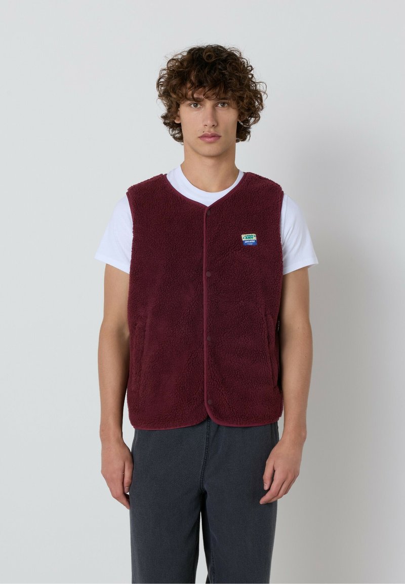 Tumši sarkans polārdzīves vestes izstrādājums ar teksturētu virsmu, noapaļotu apkakli un spiedpogu aizdari, uzvilkts virs baltas T-kreklu. Kreklā ir neliels logotips kreisajā krūtī.