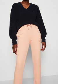 Persoon in een zwarte V-hals trui en lichtroze sweatpants met koordjes staat tegen een effen achtergrond.