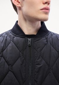 Δερμάτινη μαύρη ζακέτα bomber με ψηλό γιακά, διαθέτοντας μπροστινό φερμουάρ και ριμπ μανσέτες. Το ύφασμα έχει λεία υφή και γυαλιστερή εμφάνιση.